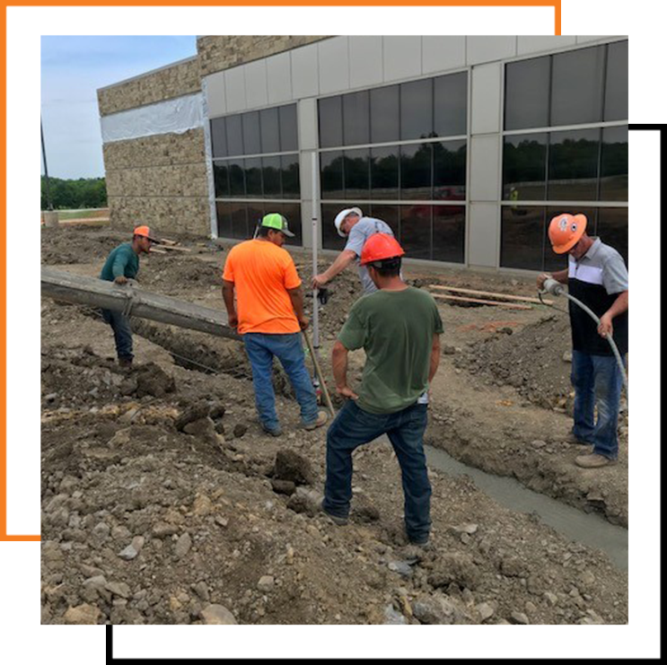 Construction workers wearing safety helmets inspect and measure a trench beside a commercial building with large reflective windows during an early phase of site work.