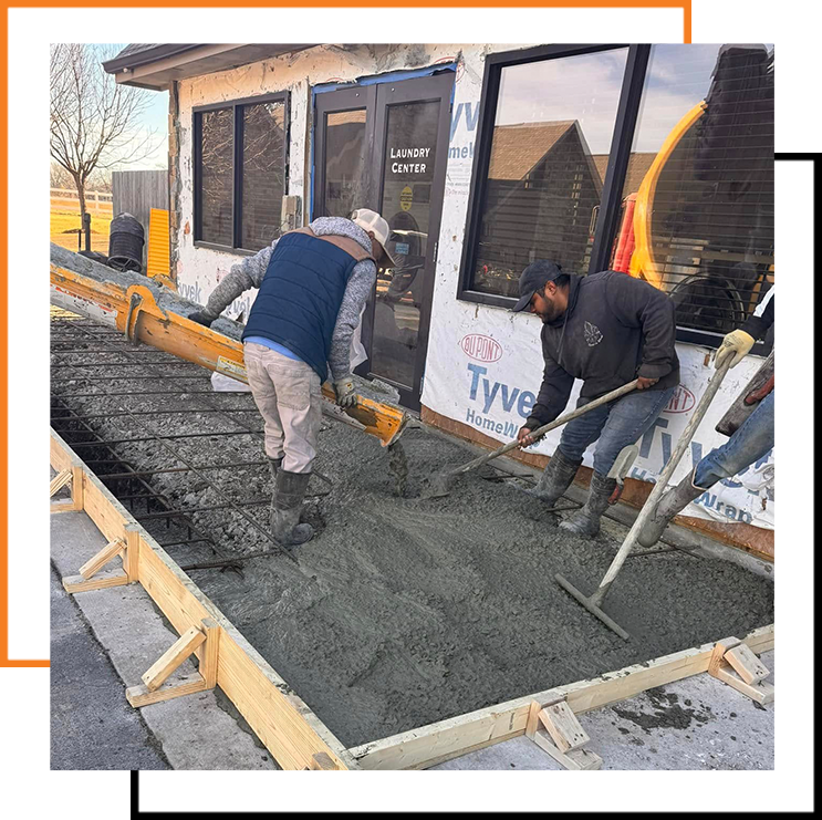 Workers pour and spread wet concrete over a rebar grid beside a building using a chute and rakes to level the surface.
